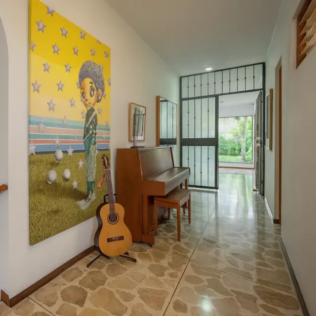 Pasillo con piso de baldosas de piedra, una pintura grande amarilla en la pared, un piano vertical de madera con su banqueta y una guitarra acústica en un soporte.