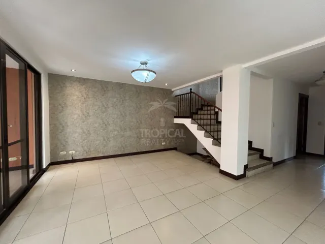 Una sala vacía con piso de baldosas claras, una pared decorativa con textura gris y una escalera con barandal de metal que sube al piso superior.