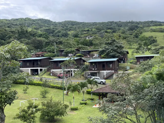 Vista exterior que muestra una hilera de cabañas de madera oscura con techos metálicos y espacios de estacionamiento, con un fondo de colinas boscosas.