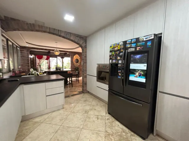 Cocina con gabinetes claros, pisos de baldosas, un horno empotrado y un refrigerador negro grande con imanes en la puerta. Un arco de ladrillo conduce a un comedor adyacente.