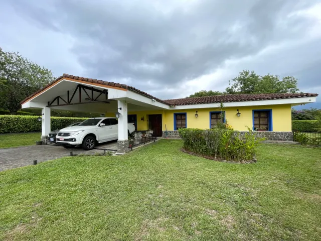 Casa amarilla de un piso con techo de tejas, marcos de ventanas azules, base de piedra y una cochera techada con dos vehículos.