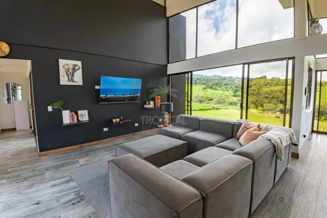 Sala de techo alto con pared de acento oscura, televisión montada en la pared, sofá modular grande y ventanales de piso a techo con vista a colinas verdes.