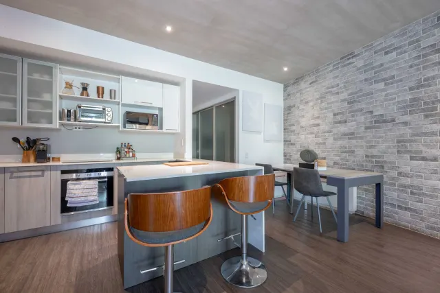 Cocina con gabinetes superiores blancos, electrodomésticos de acero inoxidable y una isla con dos taburetes de madera. Al lado hay una mesa de comedor con sillas grises junto a una pared de ladrillo gris.