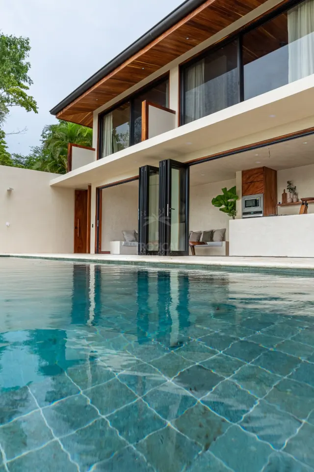 Exterior de casa de dos pisos con una piscina en primer plano, aleros de madera y puertas de vidrio plegables que abren hacia un área de estar interior.