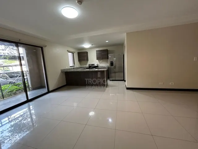 Sala abierta con pisos de cerámica brillante que conecta con una cocina equipada con gabinetes de madera oscura, una barra desayunadora y electrodomésticos de acero inoxidable. Puertas corredizas de vidrio dan hacia un patio techado.