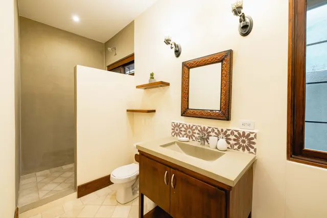Baño con un tocador de madera y lavabo de concreto, un espejo decorativo y una zona de ducha separada por un muro divisorio.