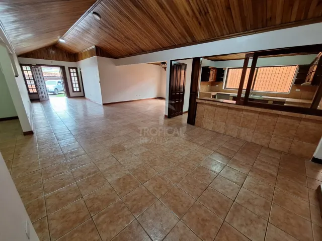 Sala vacía con piso de cerámica marrón claro, cielo raso abovedado de tablones de madera y una ventana de servicio hacia la cocina a la derecha.