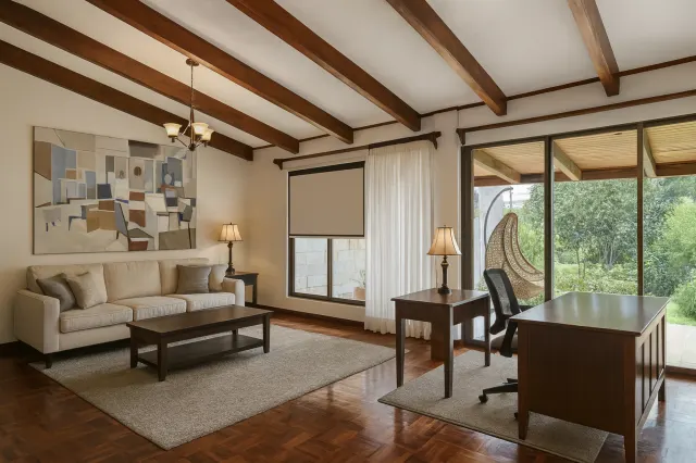 Sala de estar y espacio de trabajo con un sofá beige, escritorios de madera oscura, pisos de parquet y vigas expuestas en el cielo raso. Puertas corredizas de vidrio con vista a la vegetación.