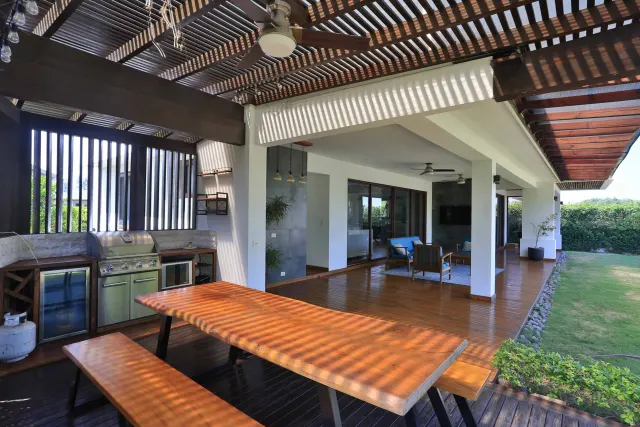 Patio exterior con mesa de comedor y bancas de madera, área de parrilla empotrada, pisos de madera y techo de pérgola. Se observa una sala en el fondo bajo el mismo techo.