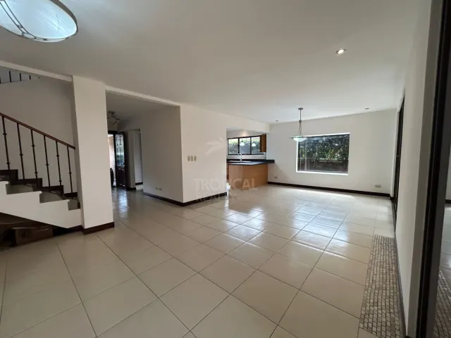 Espacio de sala vacío con pisos de cerámica claros, una escalera con barandilla oscura y vista hacia la cocina.