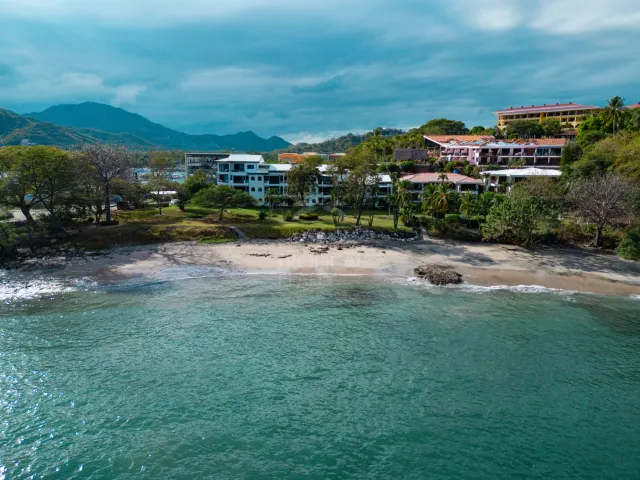 Una vista aérea que muestra edificaciones a lo largo de una playa de arena con vegetación verde, montañas al fondo y el mar en primer plano.