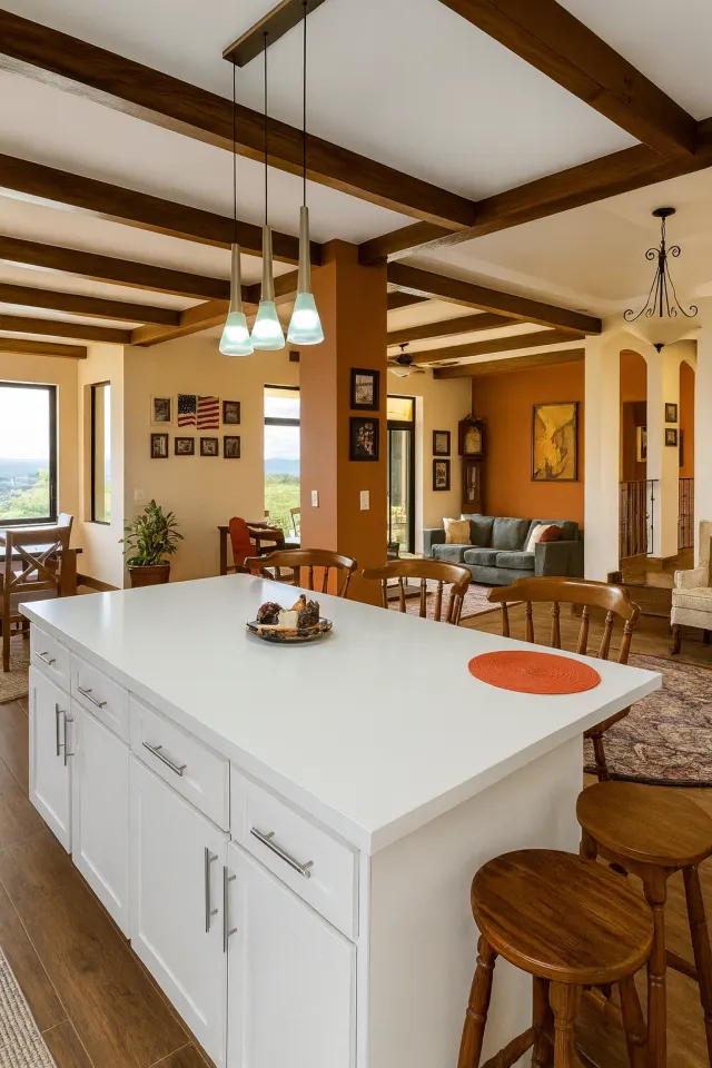 Isla de cocina con gabinetes blancos y una encimera blanca, tres lámparas colgantes y vigas de madera oscura en el techo.