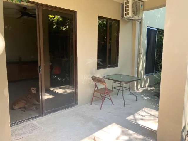 Patio techado de concreto con una mesa con cubierta de vidrio, una silla de metal y puertas corredizas de vidrio hacia el interior.